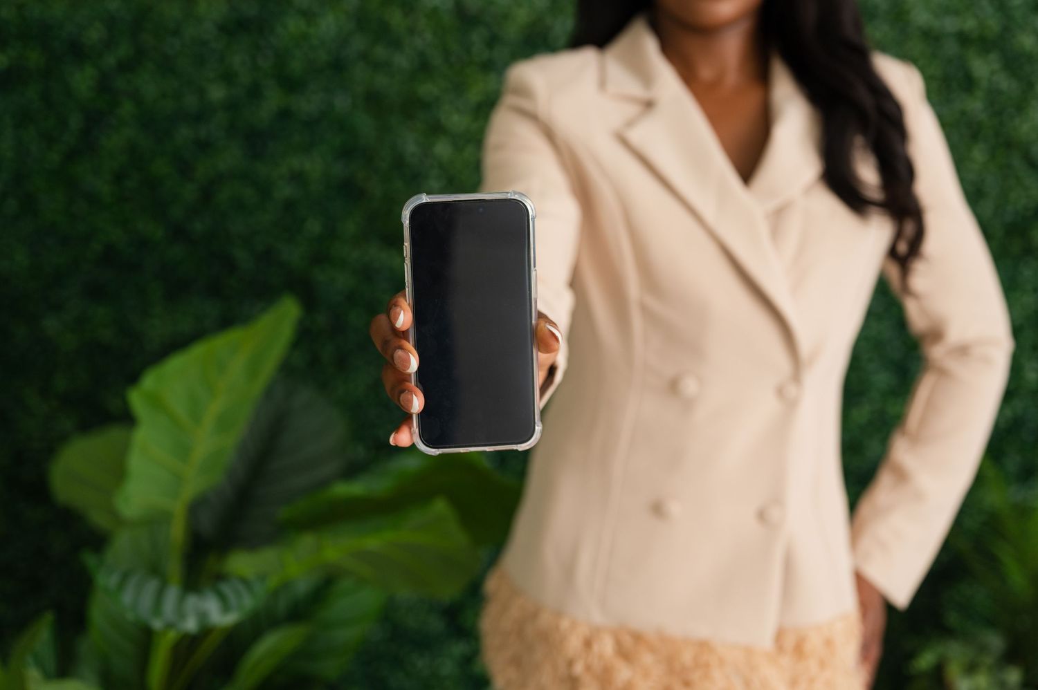 Professional in beige blazer holding up smartphone against green plant wall background.