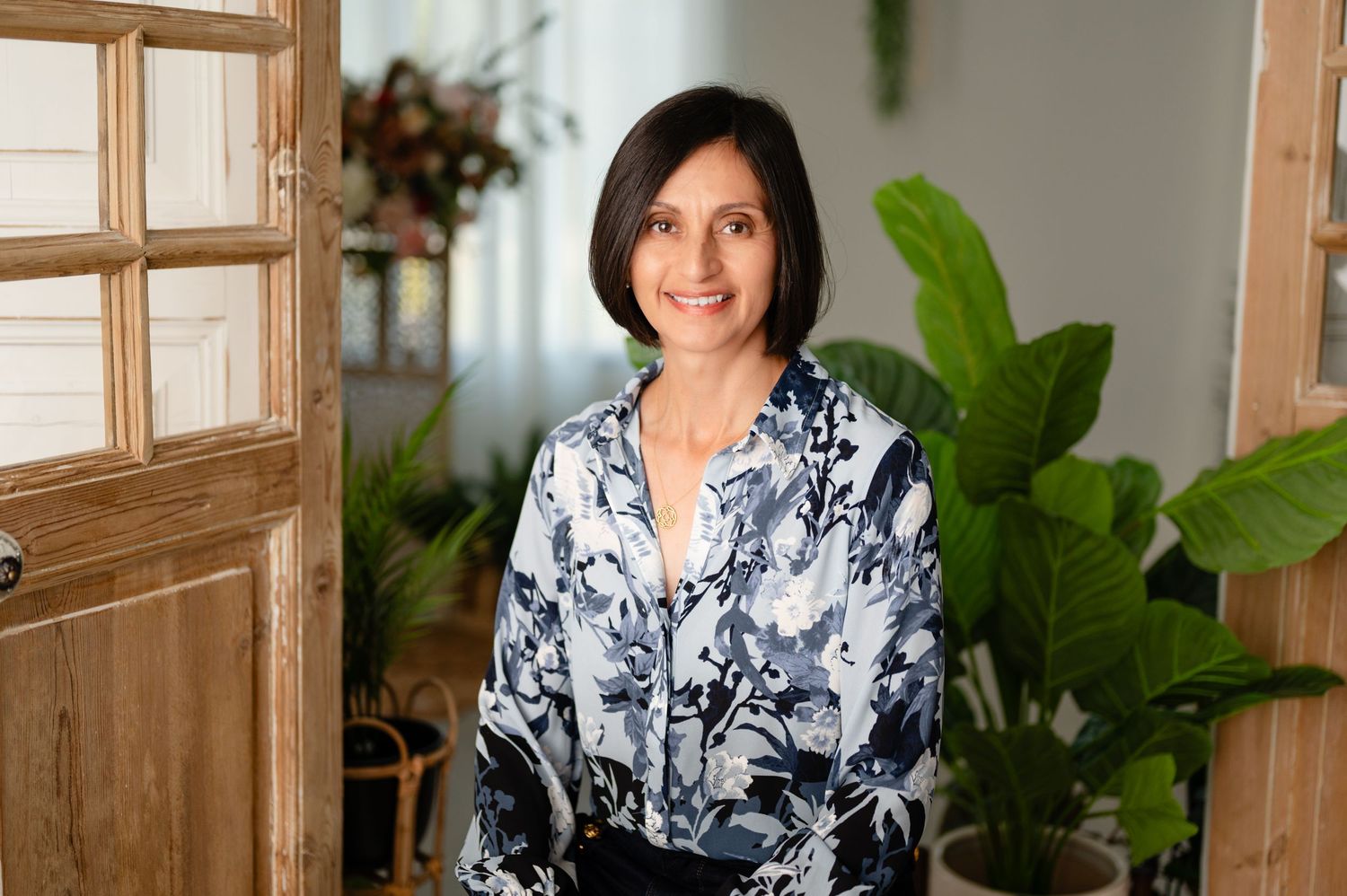 Interior portrait of woman in floral print blouse next to wooden window frame and tropical plant.