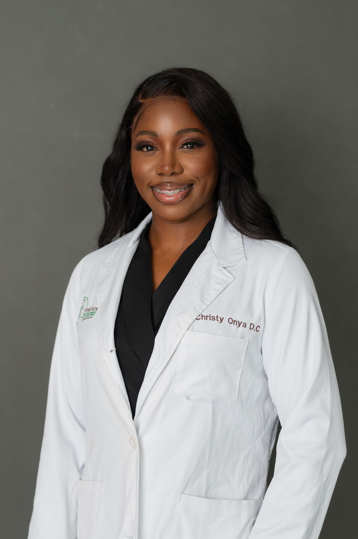 Healthcare professional in white coat and black scrubs against grey background.