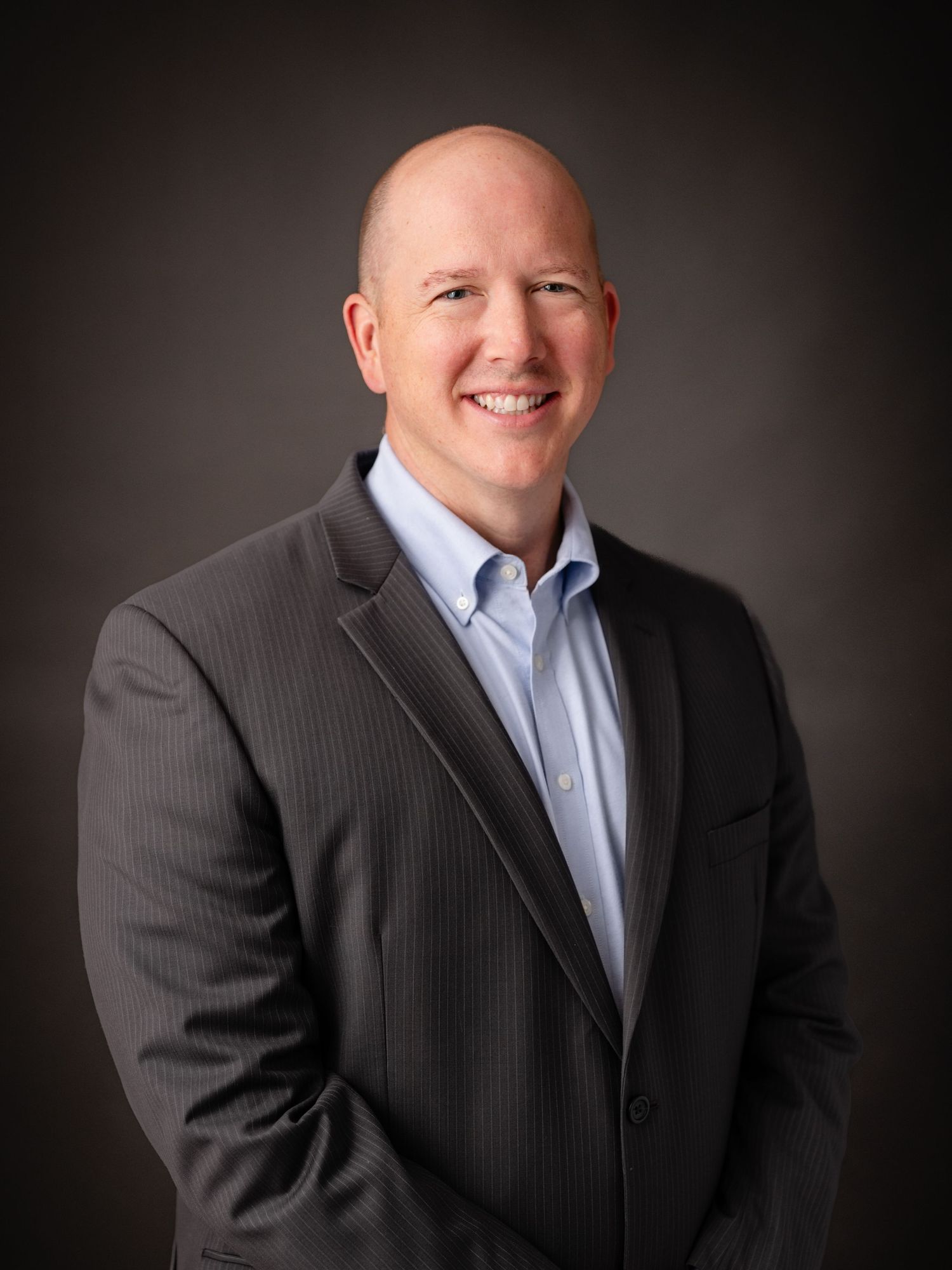 Business portrait of person wearing striped suit and light blue shirt against dark backdrop.