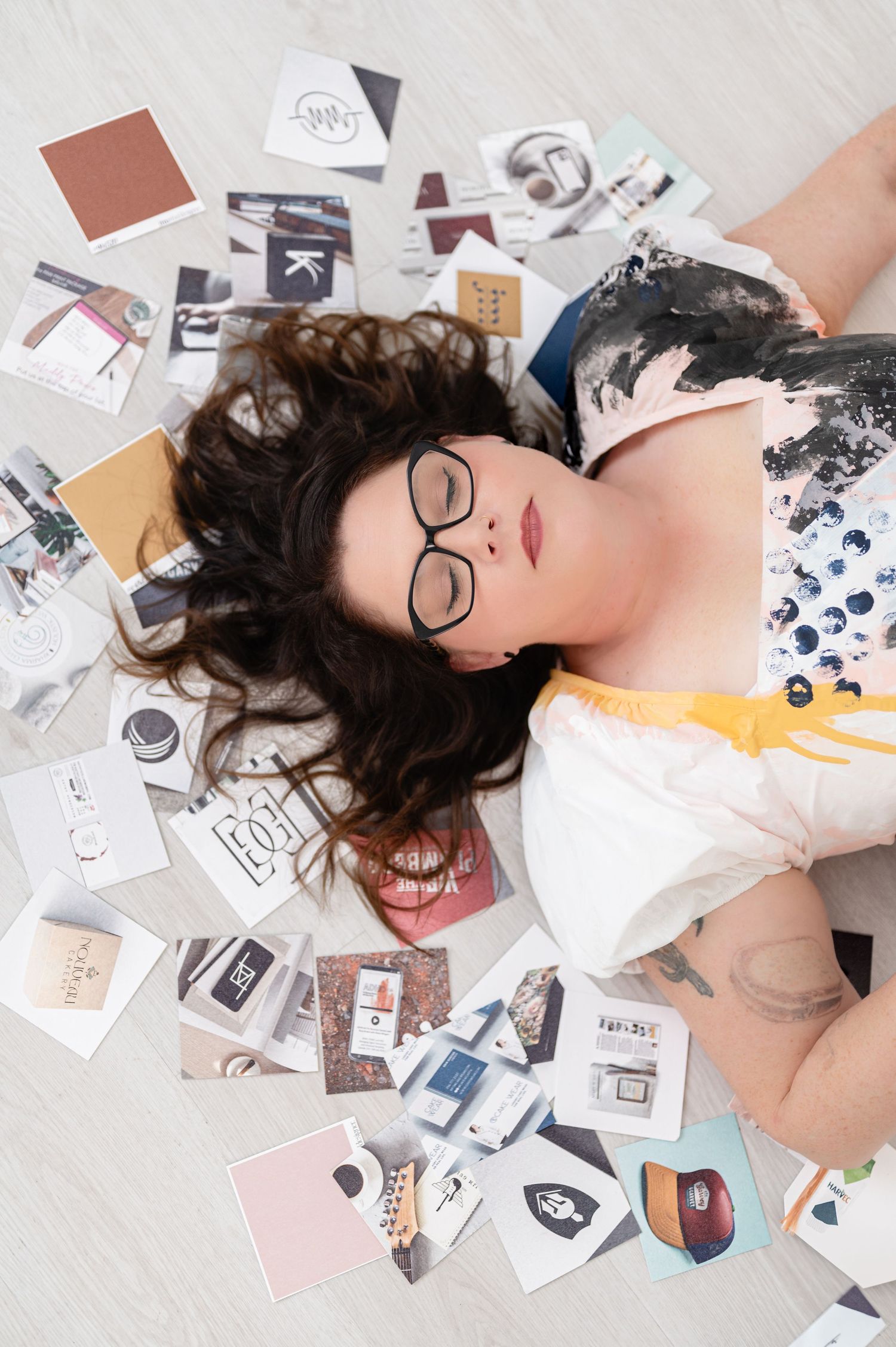Overhead view of someone wearing glasses lying on floor surrounded by scattered photographs and paper designs.