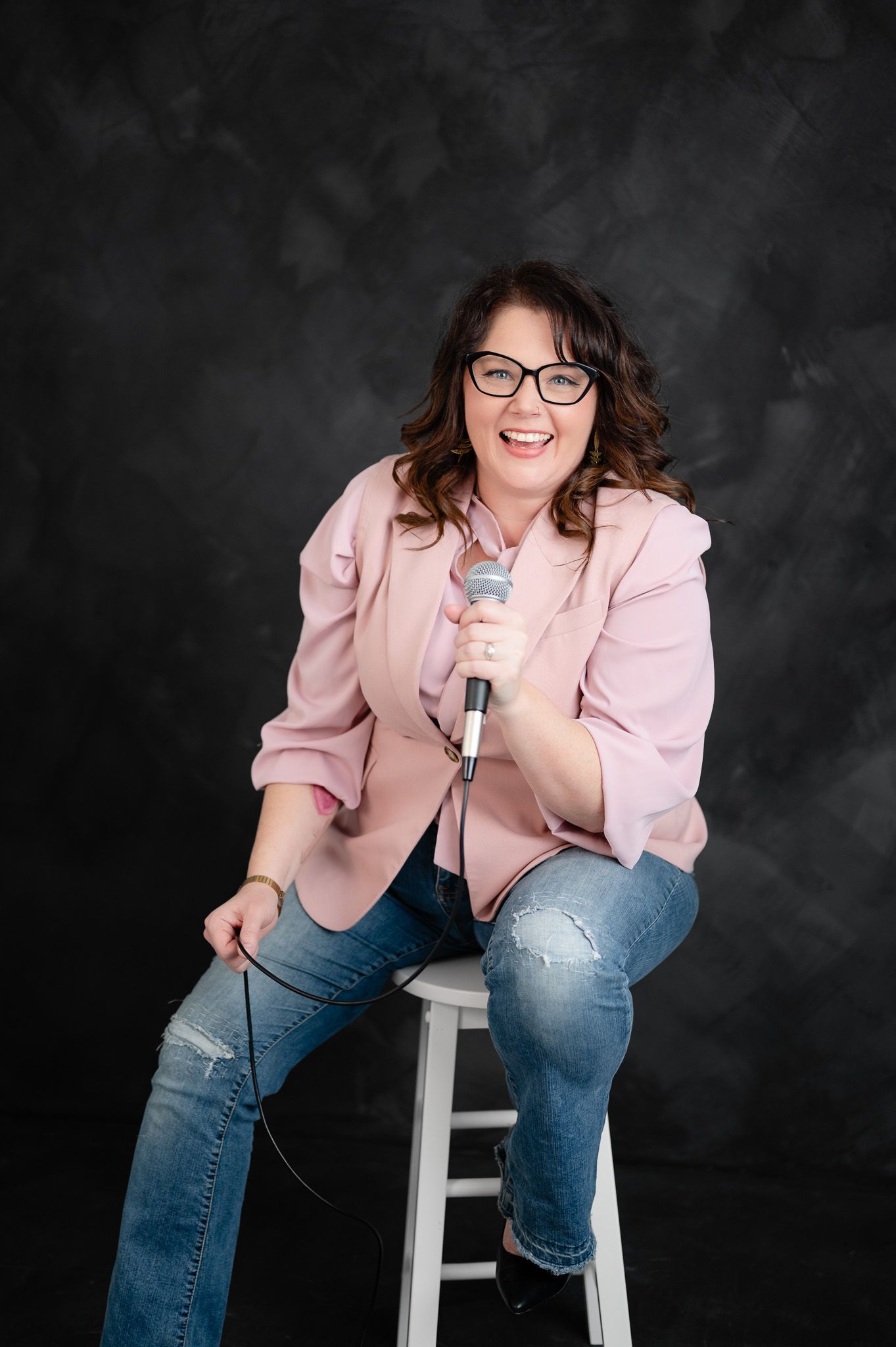 Professional portrait series showing someone in a pink blazer and jeans seated casually on a white stool against dark backdrop.