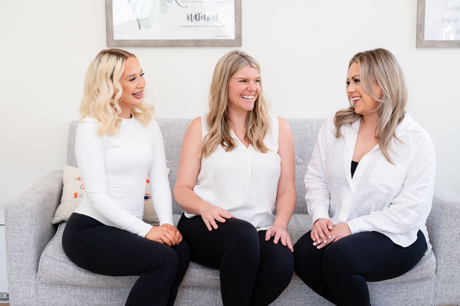 Three women in white tops sitting and laughing together on grey couch.