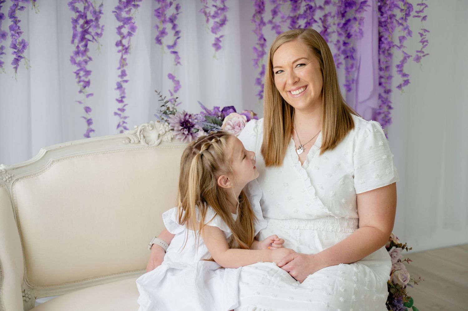 Two people in white dresses seated on an elegant cream sofa with purple floral backdrop.