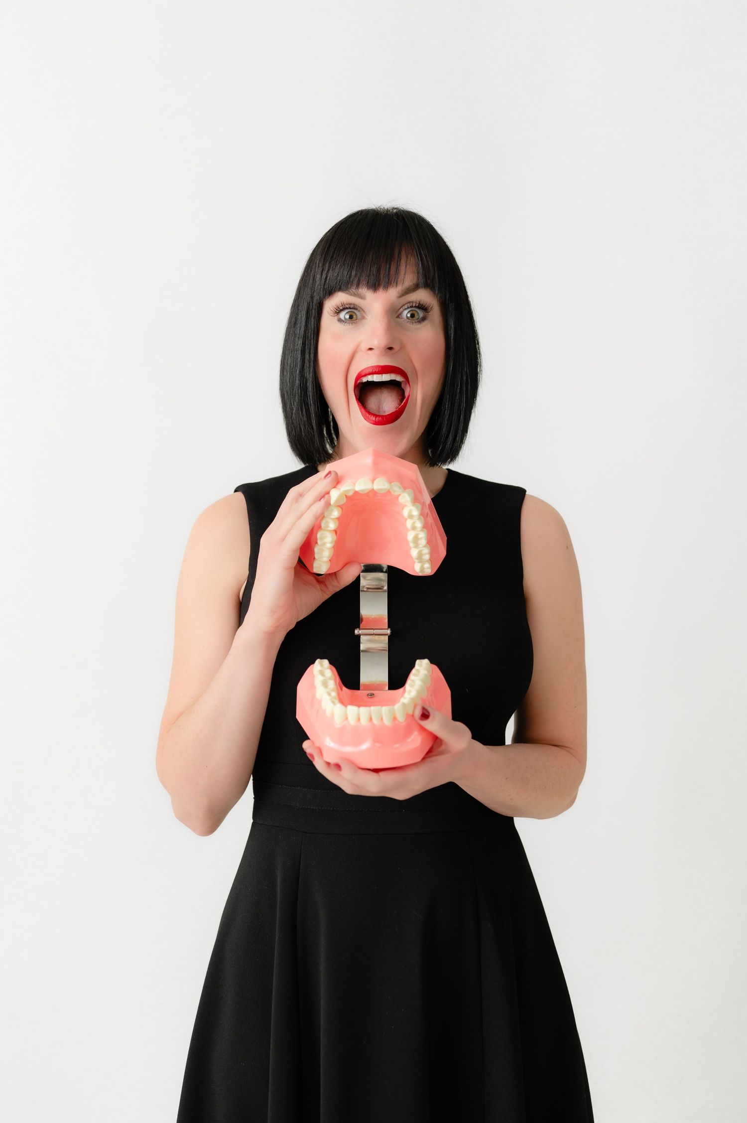 Person in black dress playfully holding oversized dental model with excited expression against white background.