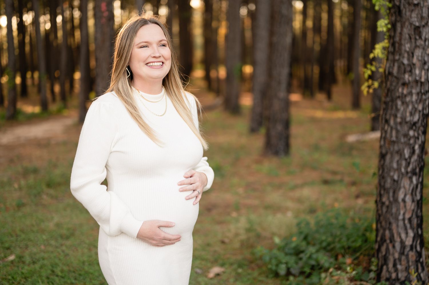 Maternity portrait in white sweater against forest background at golden hour.