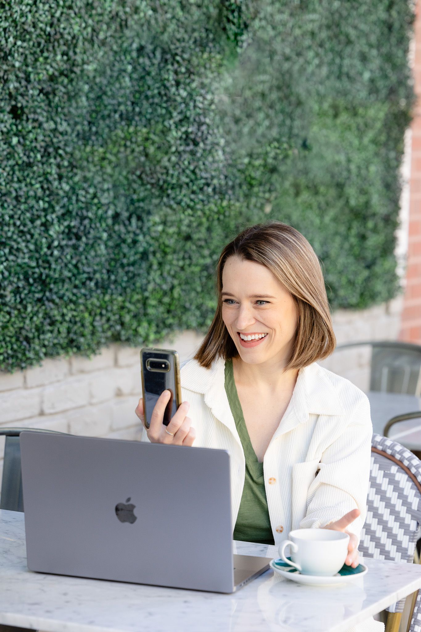 Professional working outdoors at cafe with laptop and phone against greenery wall.