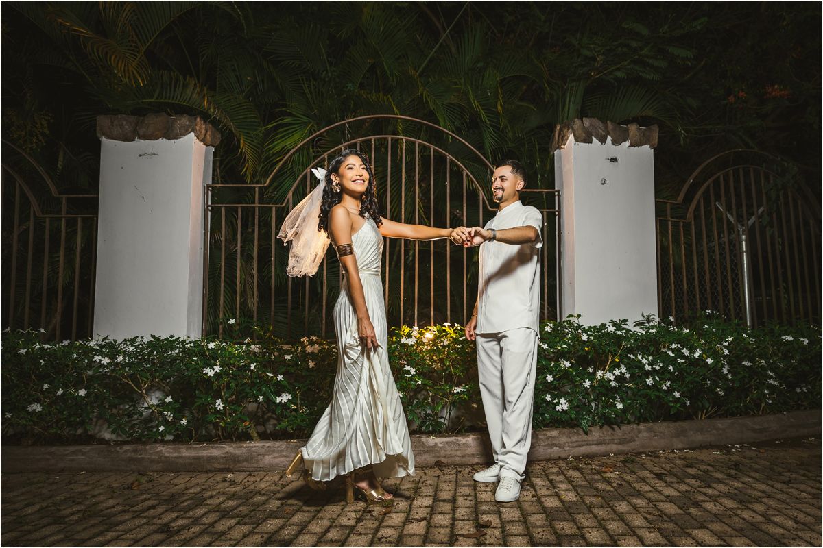 A couple in elegant wedding attire dances and poses together on a cobblestone path at night.