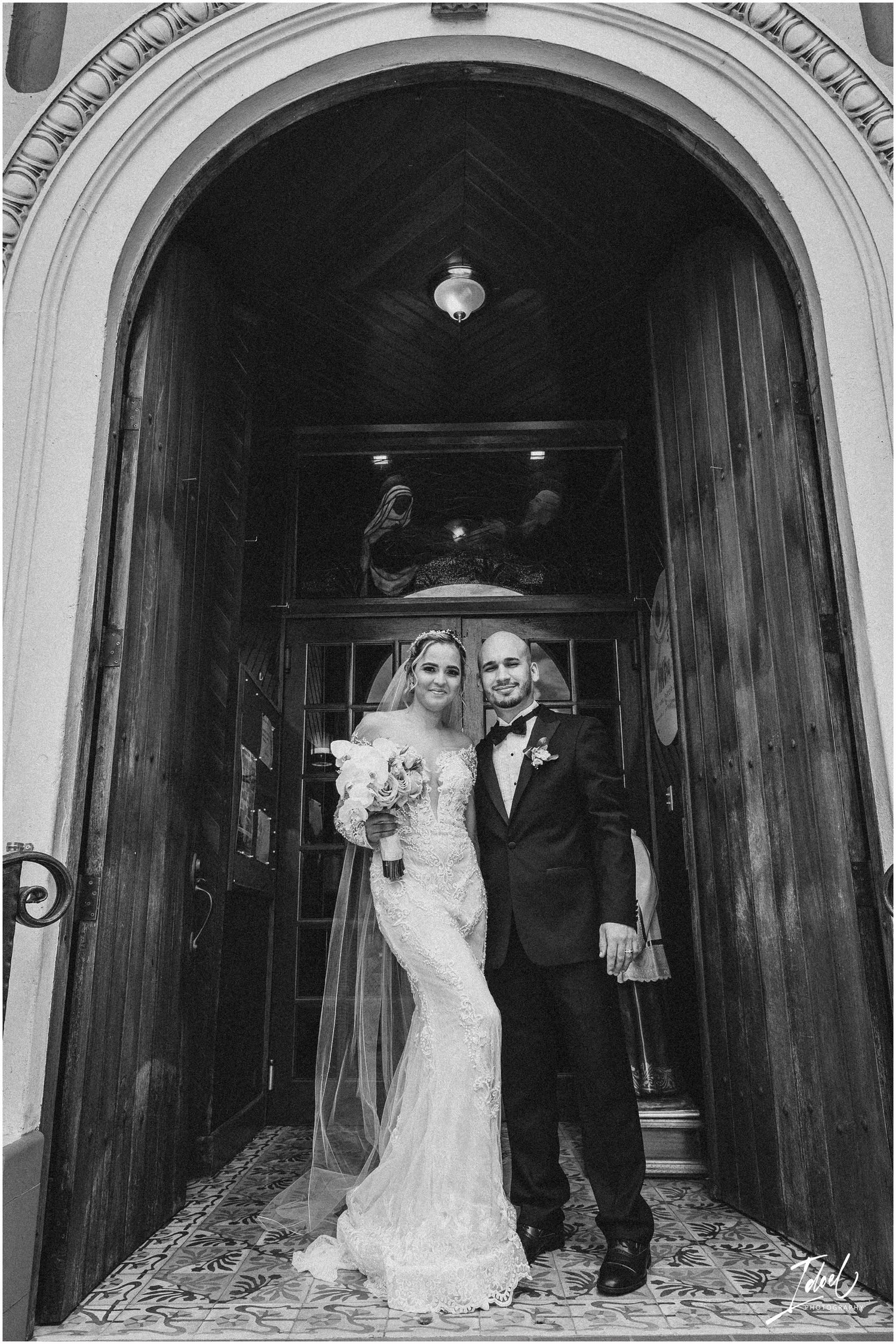 A black and white portrait of newlyweds standing under a grand Gothic archway entrance.