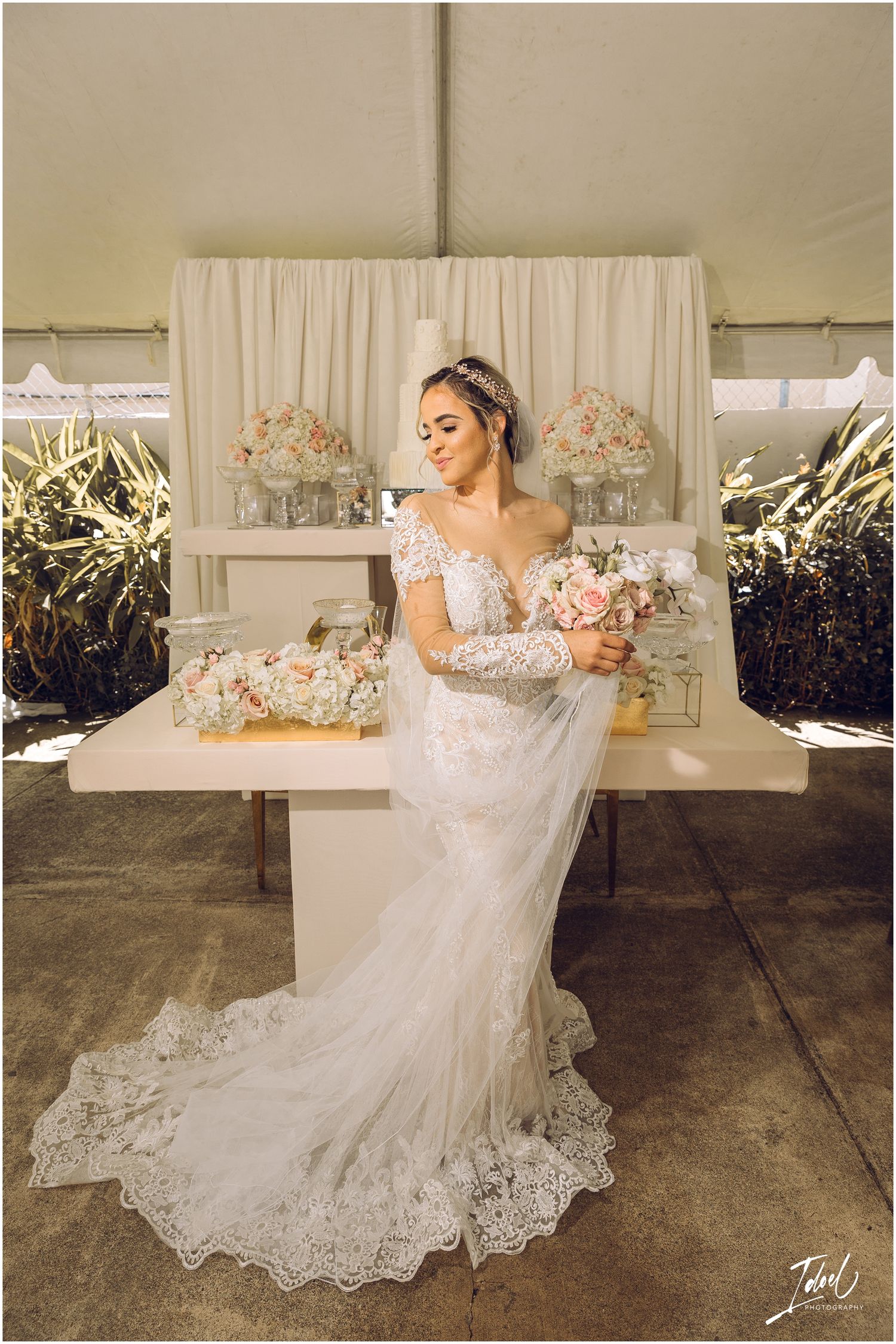 Bride in detailed lace gown poses with pink bouquet against beige backdrop and floral arrangements.