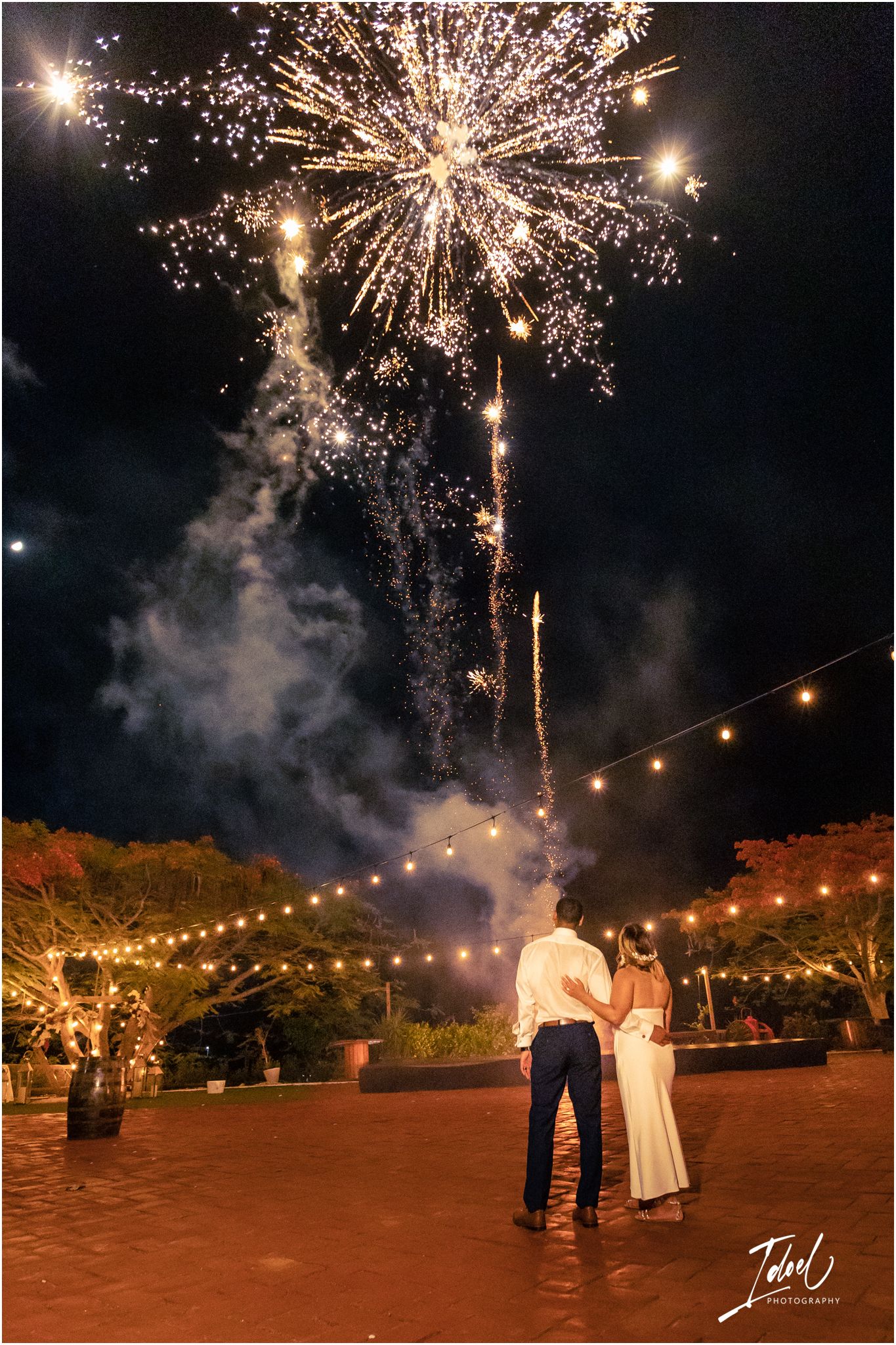 Golden fireworks explode over an outdoor wedding reception with string lights.