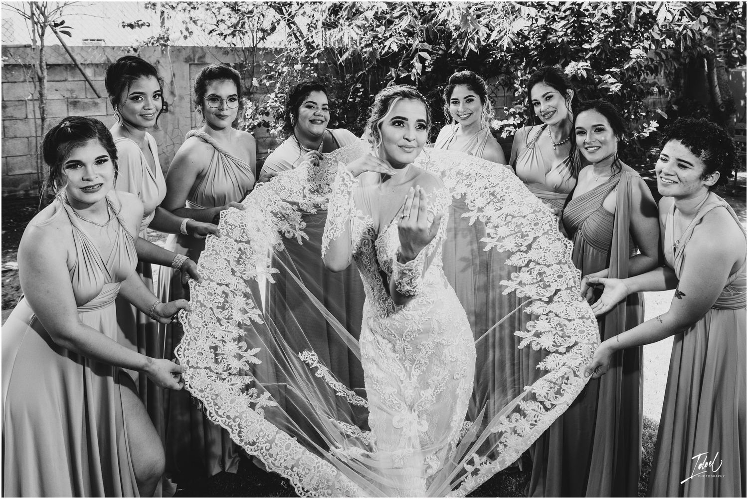 Black and white photo of bridesmaids holding bride's veil in dramatic outdoor portrait.
