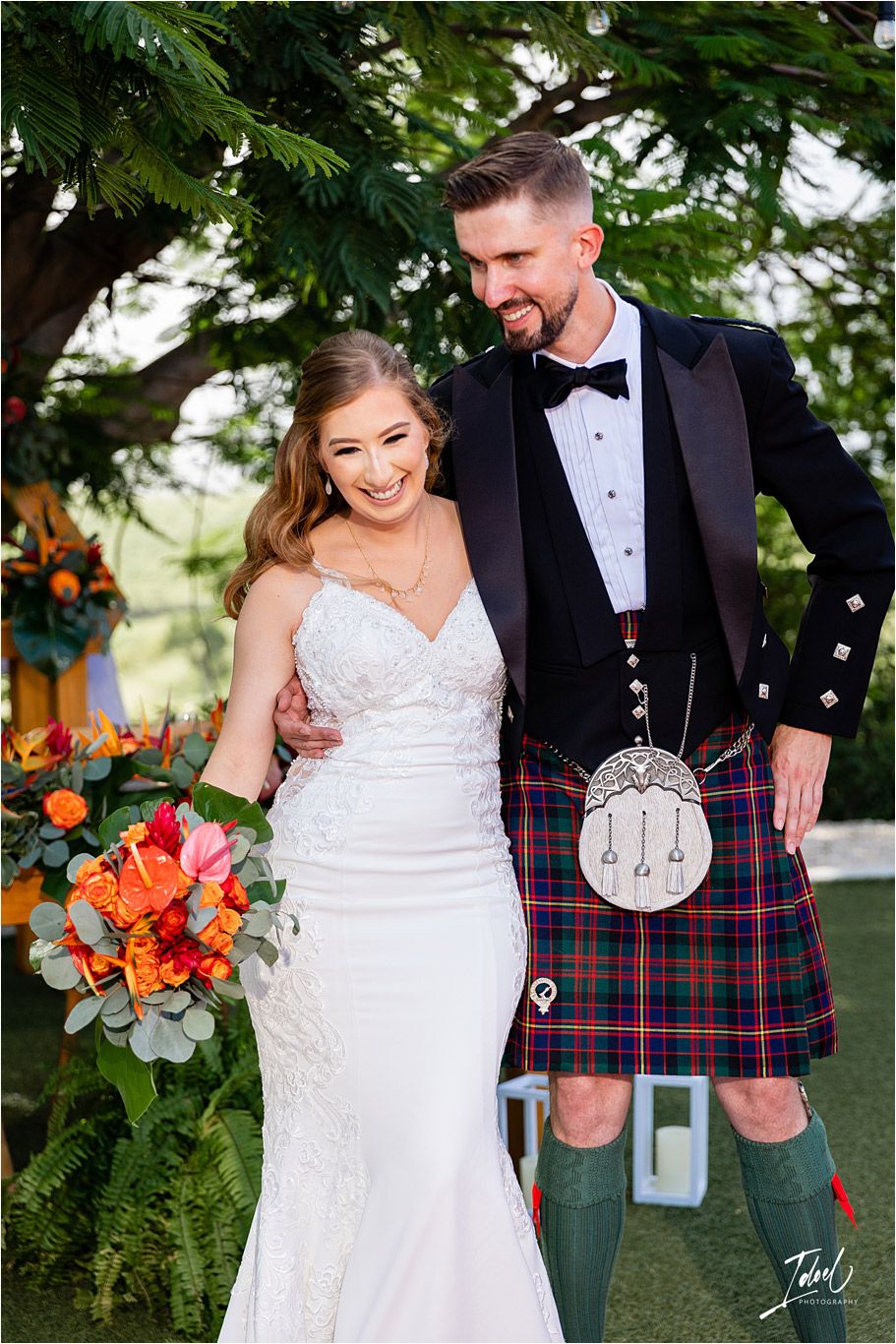 Wedding couple in tropical garden setting with bride holding orange bouquet and groom in full Scottish dress.