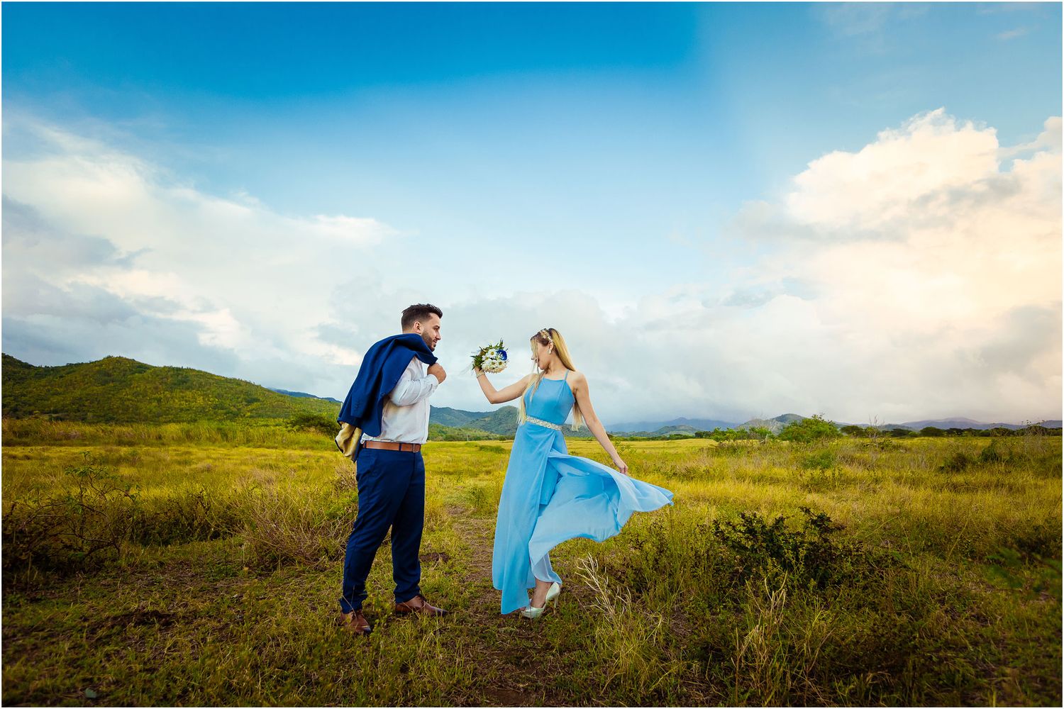 Two people in formal wear dance playfully in a golden meadow with mountains on the horizon.