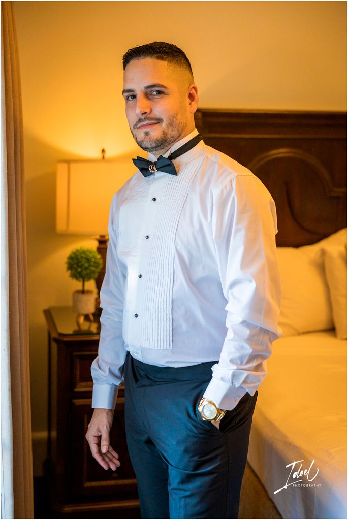 Elegant formalwear attire featuring a white pleated tuxedo shirt with black bow tie and navy slacks in a warm-lit bedroom.
