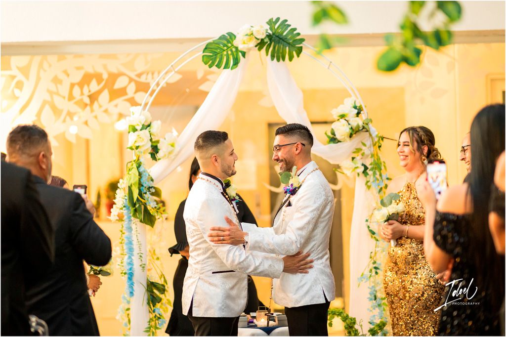 Two people share an intimate moment during their wedding ceremony under elegant floral decor and golden lighting.