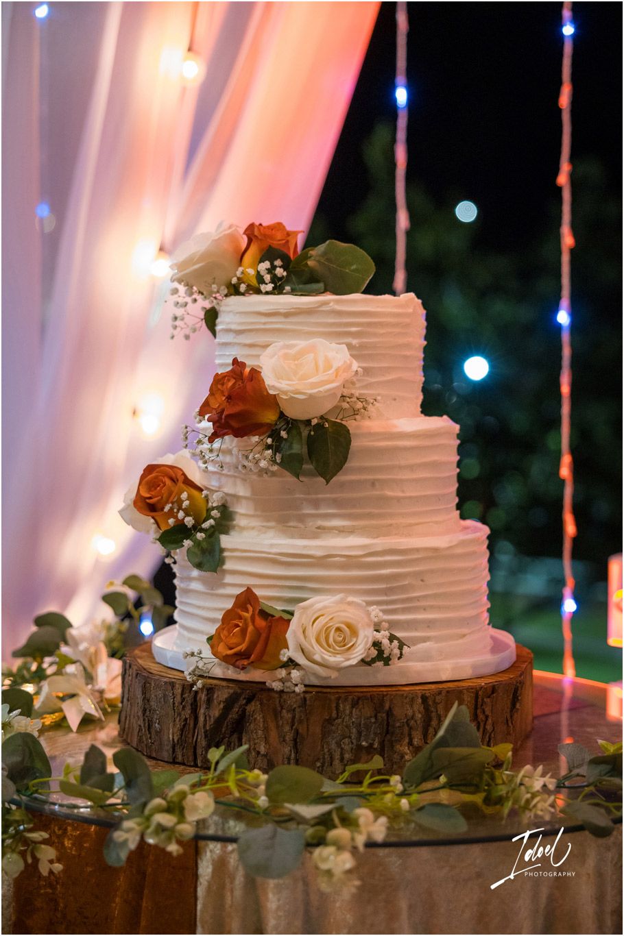 White textured wedding cake decorated with roses sits on rustic wood slice stand with soft pink lighting in background.