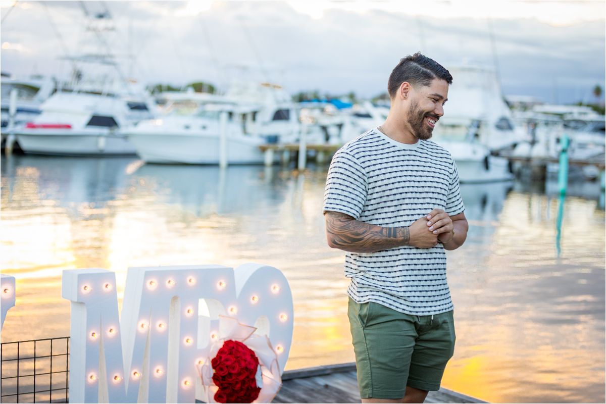 Person in striped shirt stands by marina at sunset with illuminated marquee letters and red rose in foreground.