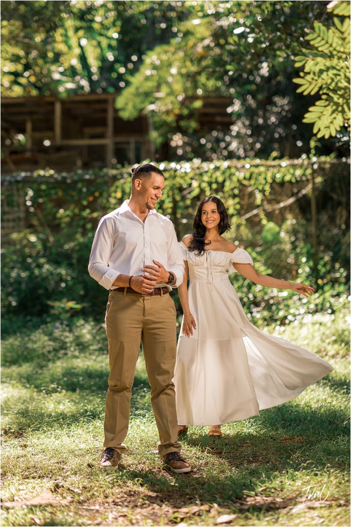 A couple dances together in a sunlit garden setting, the woman's flowing white dress swaying with their movement.
