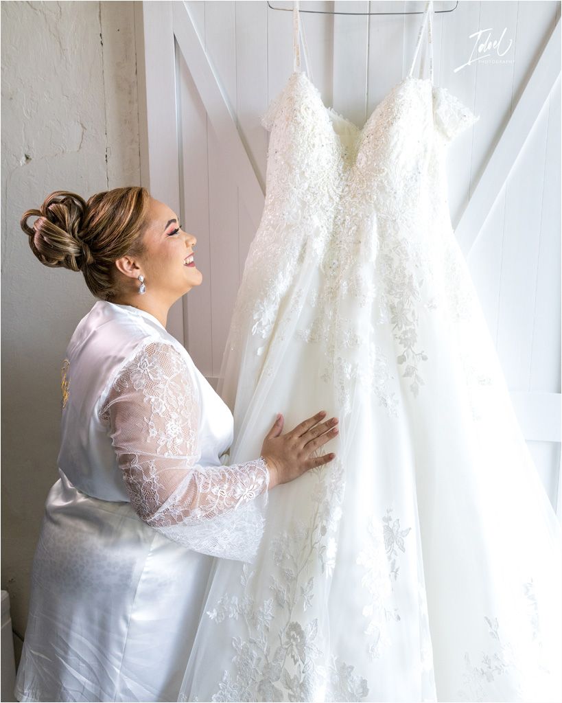 A person in a white robe admires a hanging wedding dress against a white door.