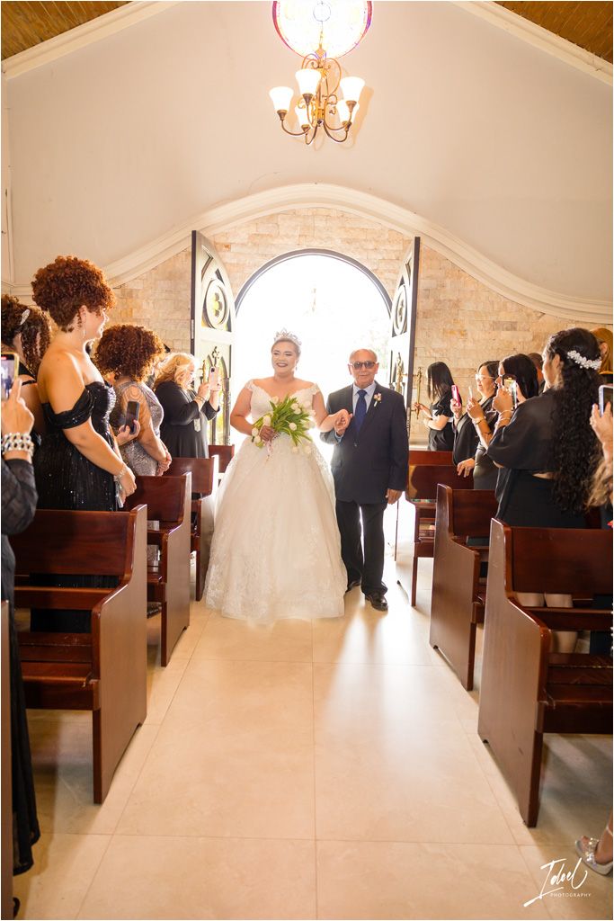 Wedding ceremony processional in a white chapel with guests lining the pews.