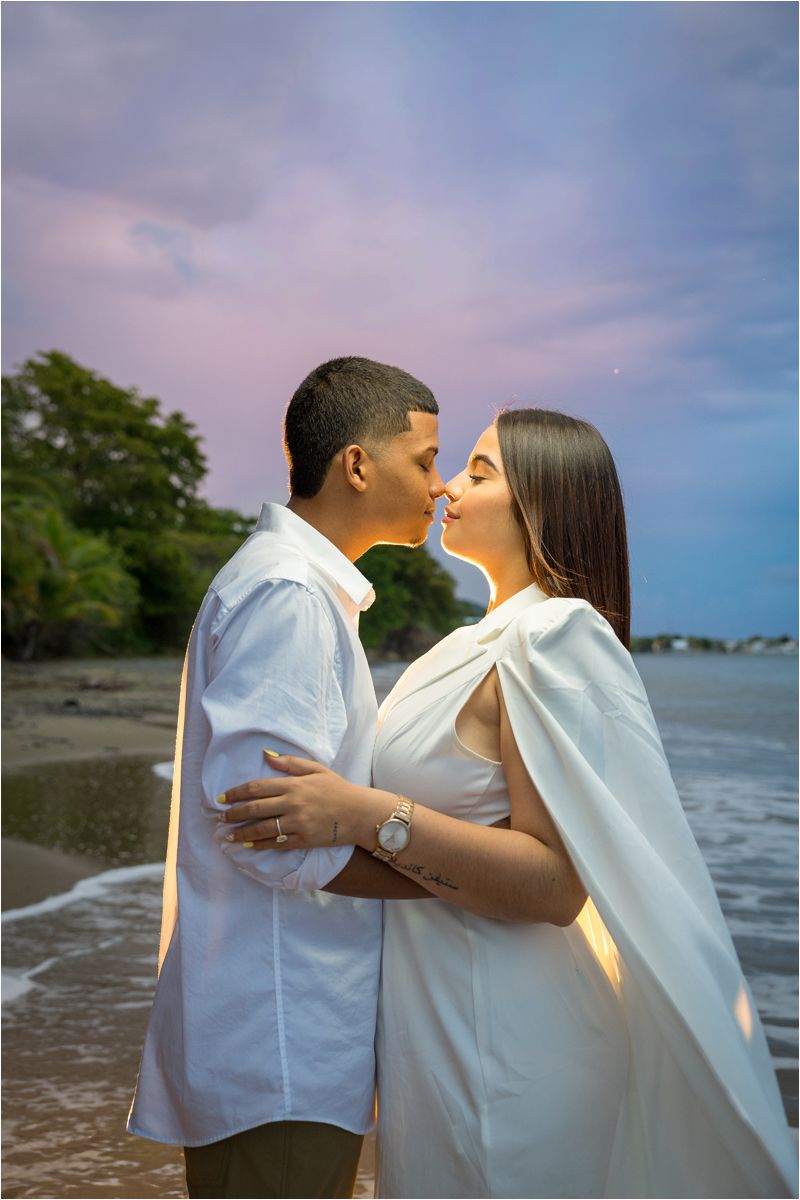 Couple in white outfits sharing a romantic moment on a beach at sunset with colorful sky.