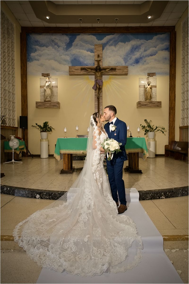 Bride and groom share a kiss at the altar of a Catholic church, with a large wooden cross and mural backdrop.