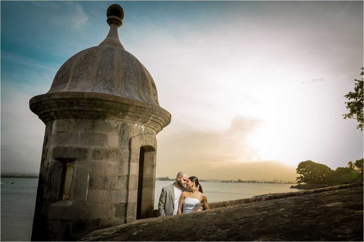A romantic couple embraces at sunset near a stone lighthouse overlooking the ocean.