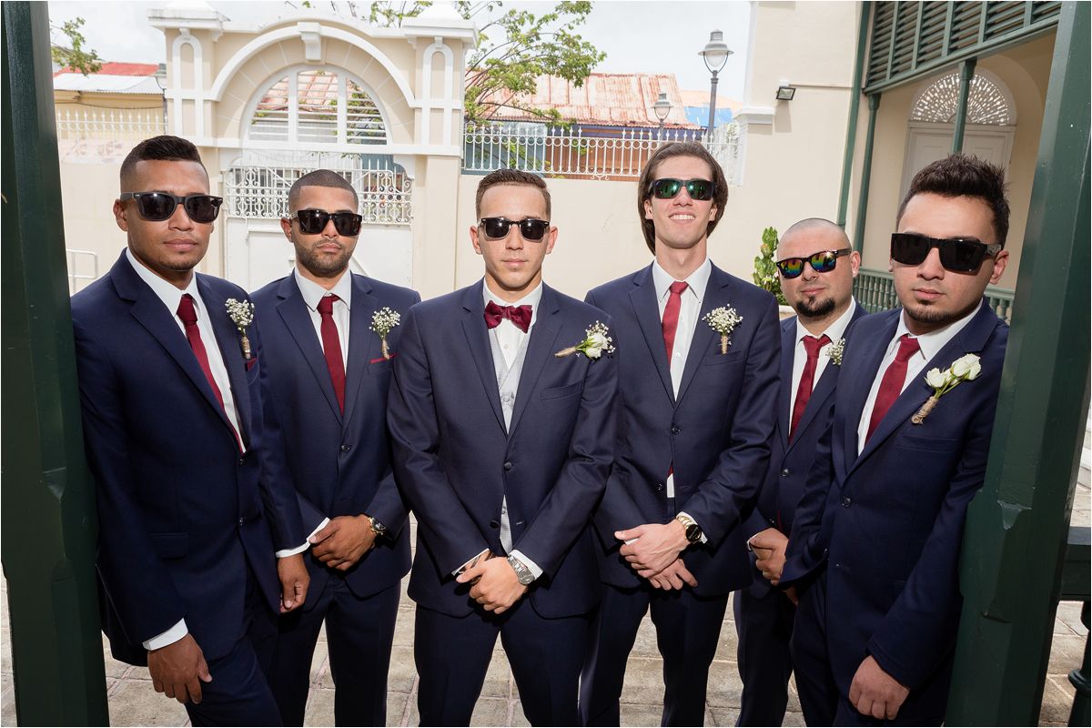 Groomsmen in matching navy suits and red ties pose together wearing sunglasses outside an ornate venue entrance.