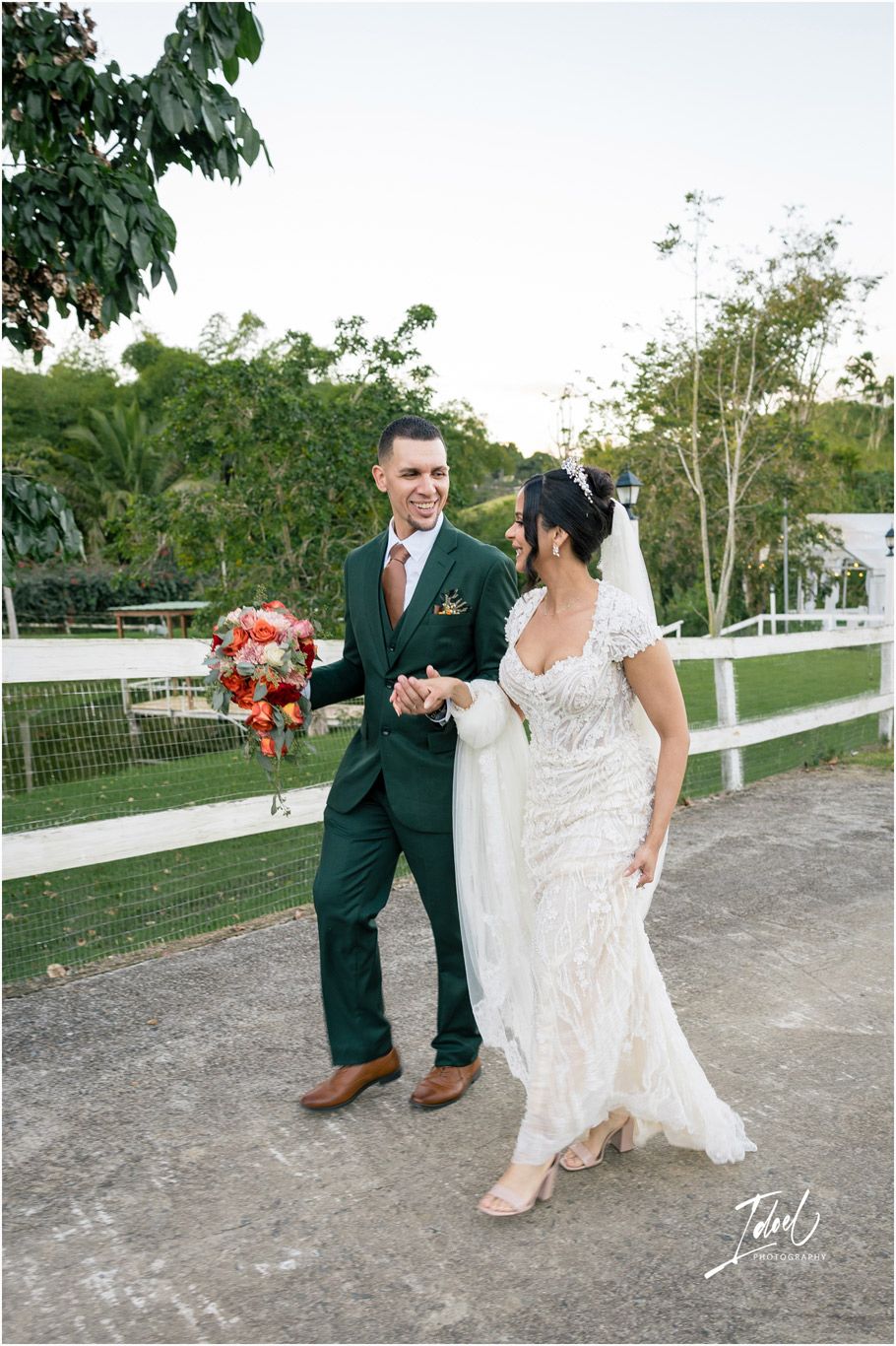 A couple walks along a pathway with white fences at an outdoor wedding venue, the bride holding a coral bouquet.