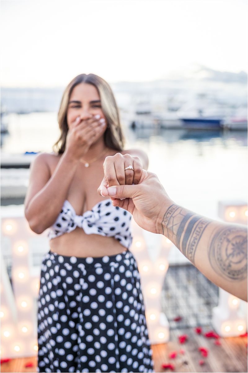 A romantic proposal moment captured at a marina with a polka dot outfit and rose petals in the background.