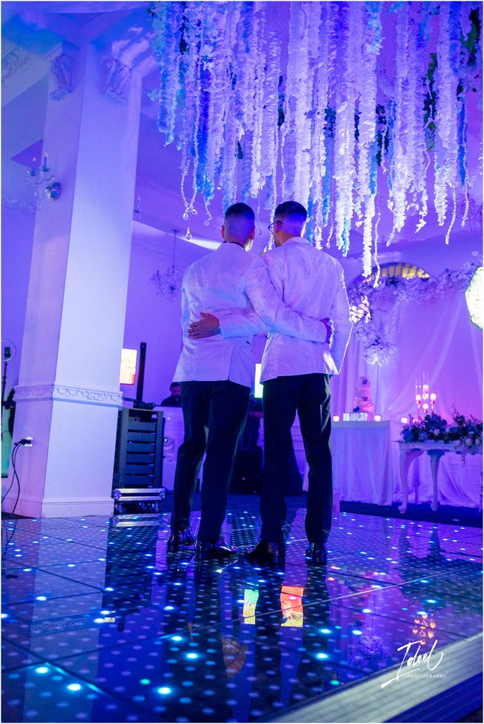 Two people in white shirts embrace on a dance floor illuminated by purple lighting and hanging crystal decorations.