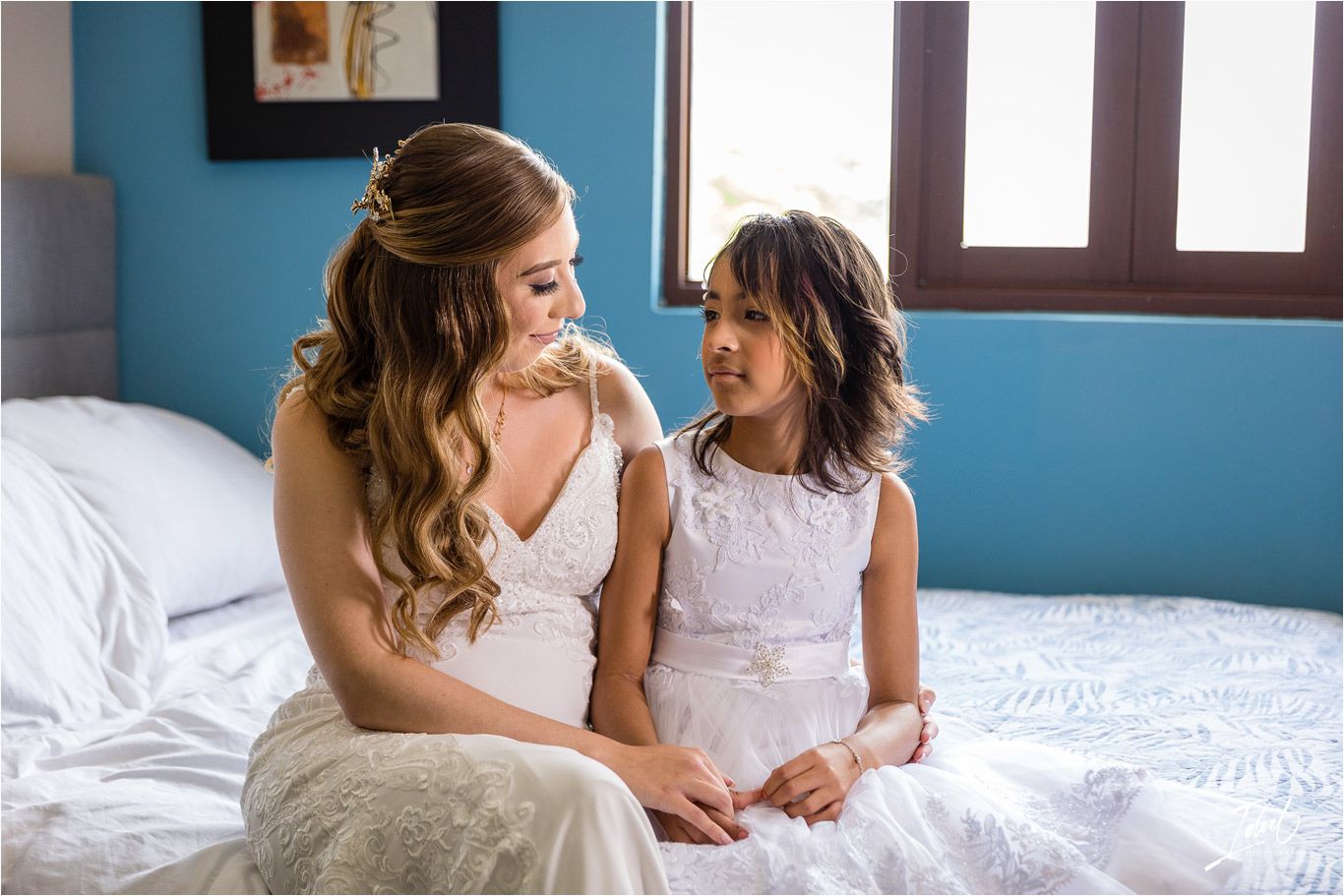 Heartwarming moment between bride and young girl in white dresses sitting on bed.