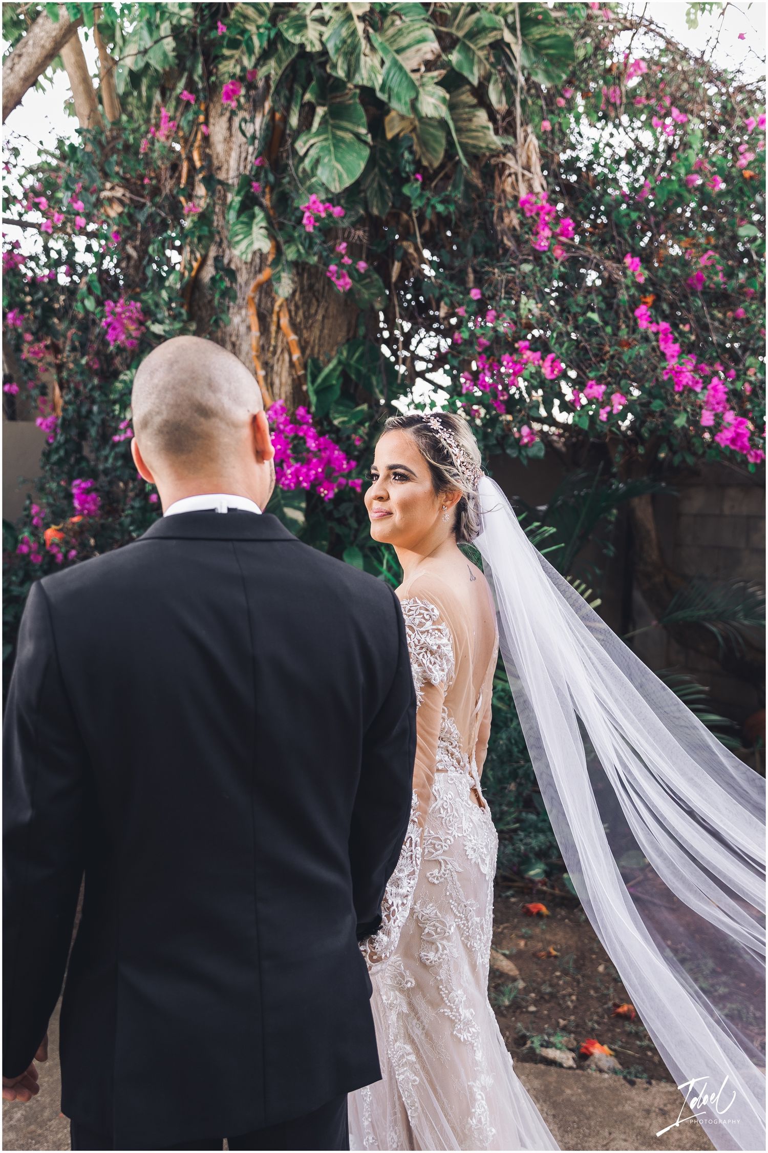 A bride in a lace wedding dress with flowing veil poses against a backdrop of magenta flowers.