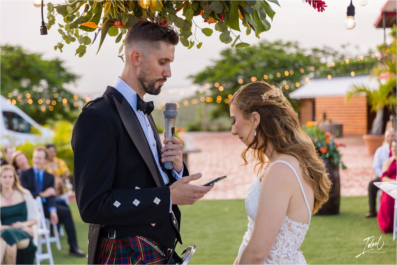 A touching moment during a wedding ceremony as a couple shares their vows under decorative lighting and greenery.