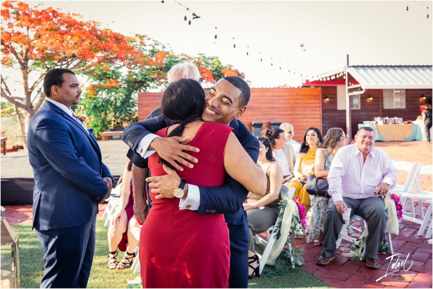 Emotional embraces at outdoor wedding ceremony under vibrant orange flowering tree.