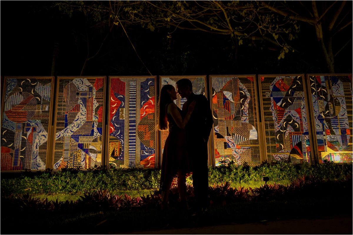 Couple silhouetted against vibrant illuminated mosaic artwork panels in a romantic nighttime setting.