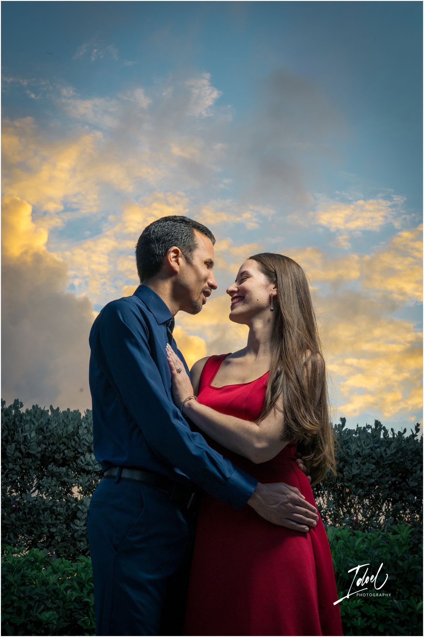 Couple in romantic embrace under dramatic sunset sky, woman in red dress and man in blue shirt sharing an intimate moment outdoors.
