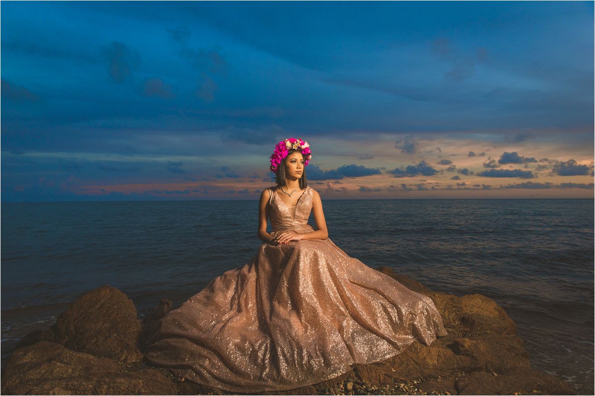 A person in a flowing champagne gown and floral crown sits on coastal rocks at sunset against a dramatic blue sky.