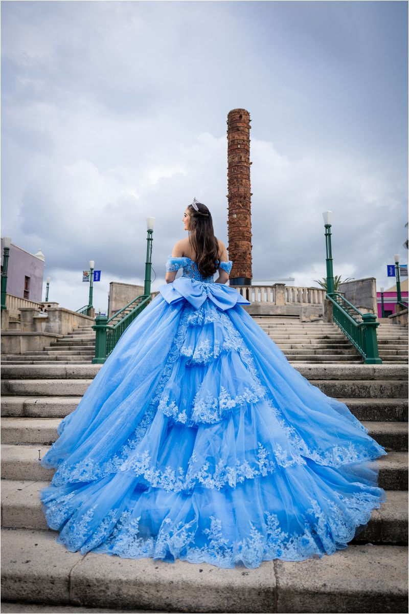 A stunning blue ball gown with white lace details flows dramatically across stone steps against a cloudy sky.