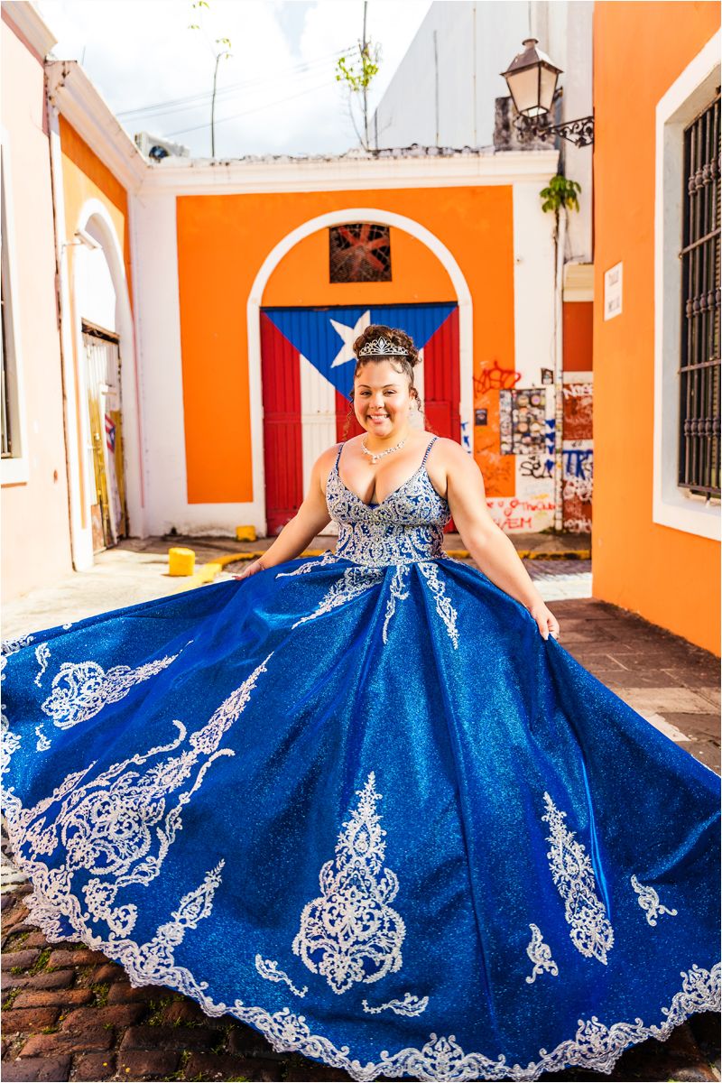 Royal blue quinceañera dress with intricate white lace embroidery displayed in a Spanish colonial style courtyard.