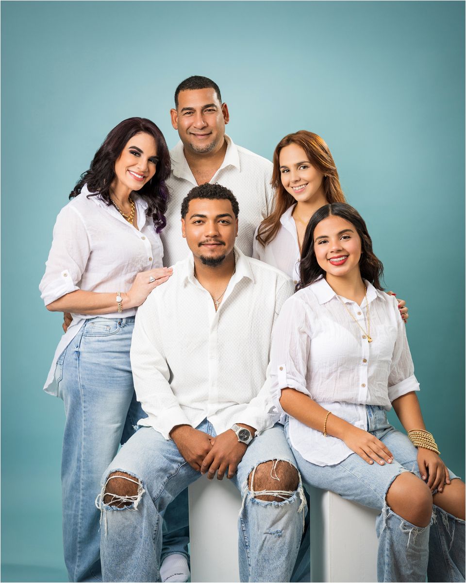 A group in white shirts and distressed jeans pose together against a turquoise studio backdrop.