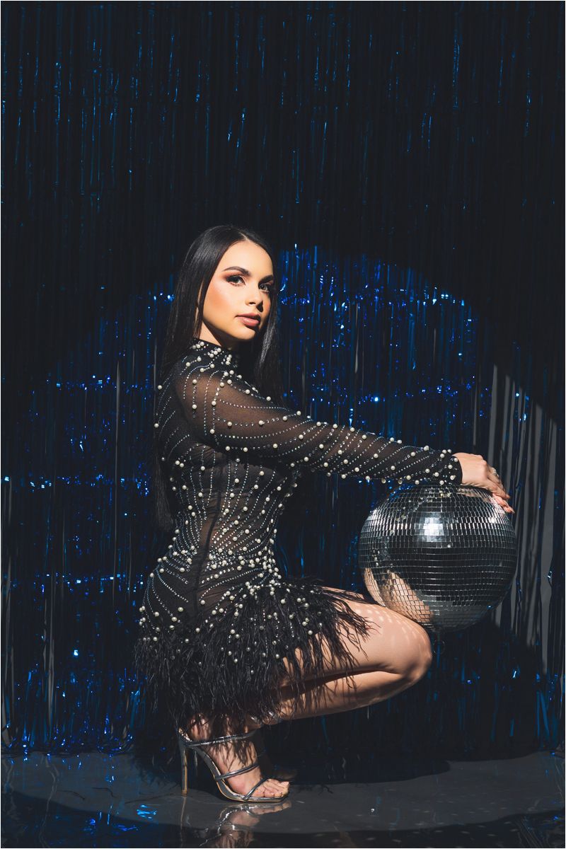 A performer in a sparkling black outfit poses dramatically against a backdrop of blue lights in a dark studio setting.
