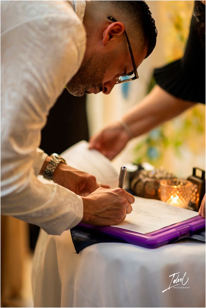 A person wearing glasses and a white dress shirt signs a document at a candlelit table.