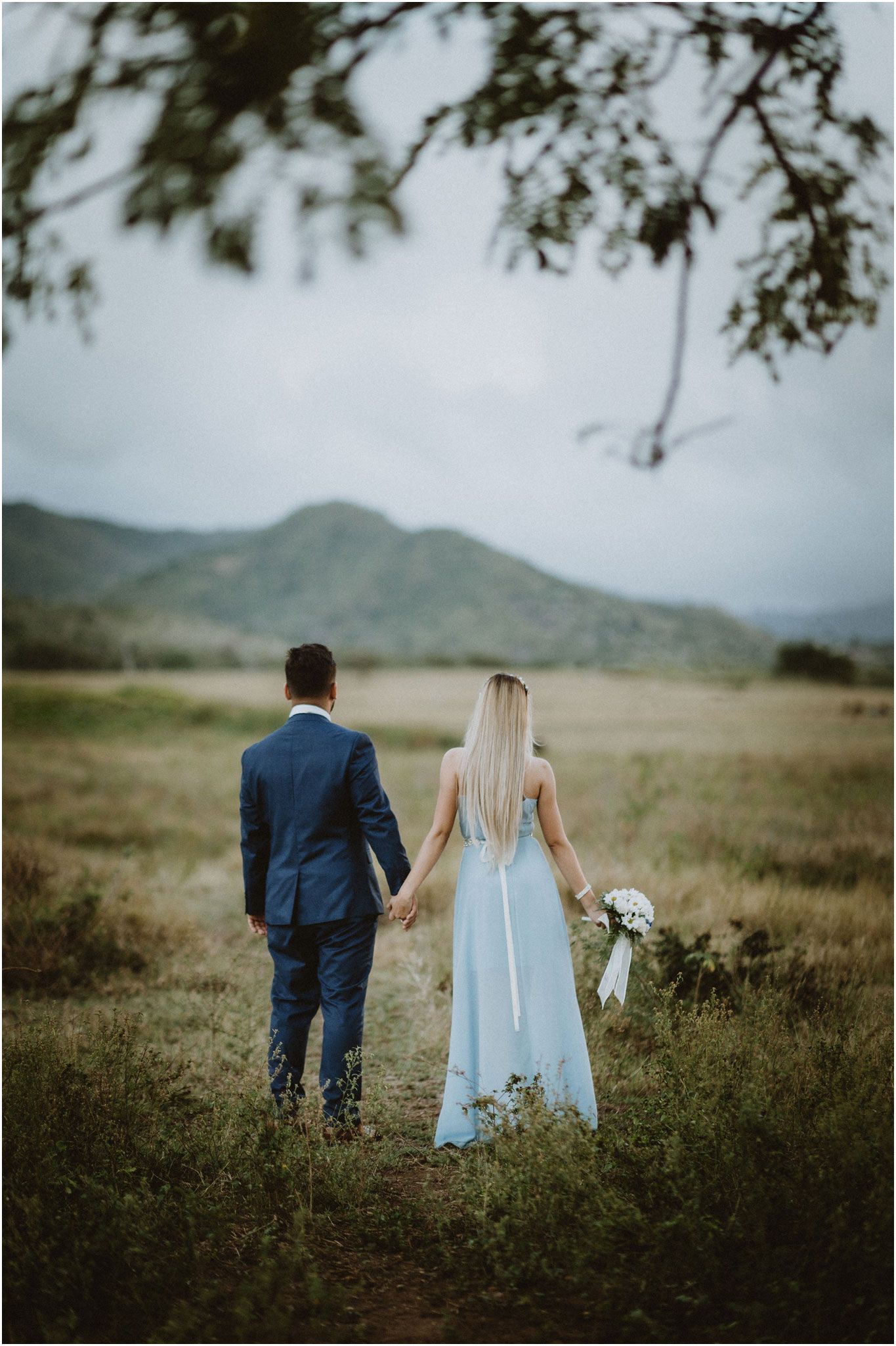 A couple walking hand in hand through a meadow with mountains in the distance.