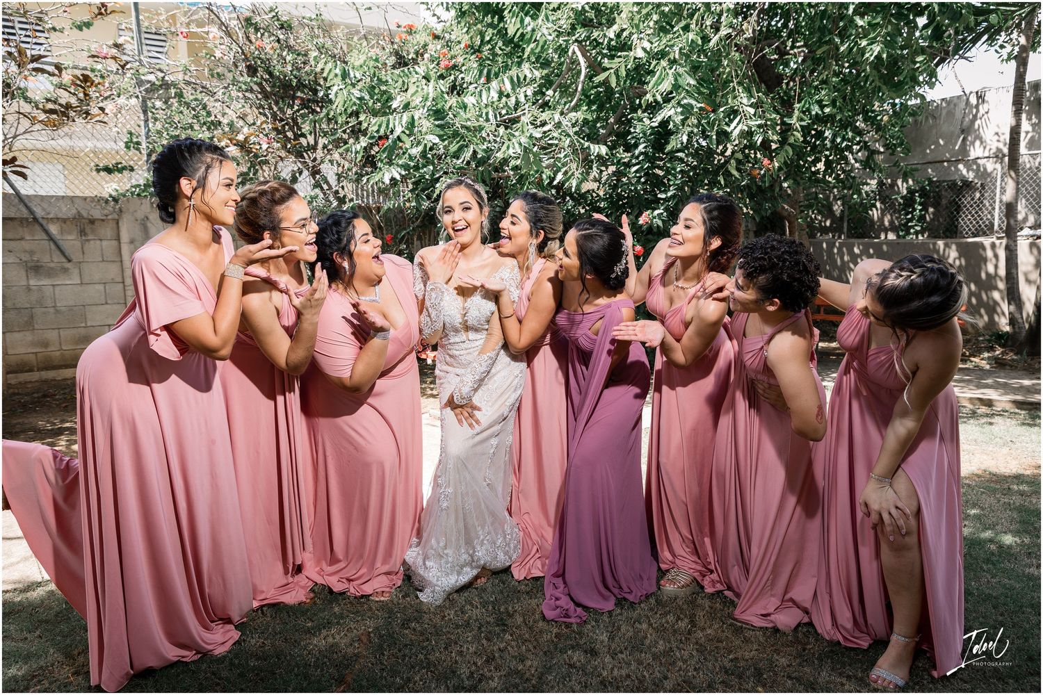 Bridesmaids in pink and purple dresses surround bride in white gown for garden photo.
