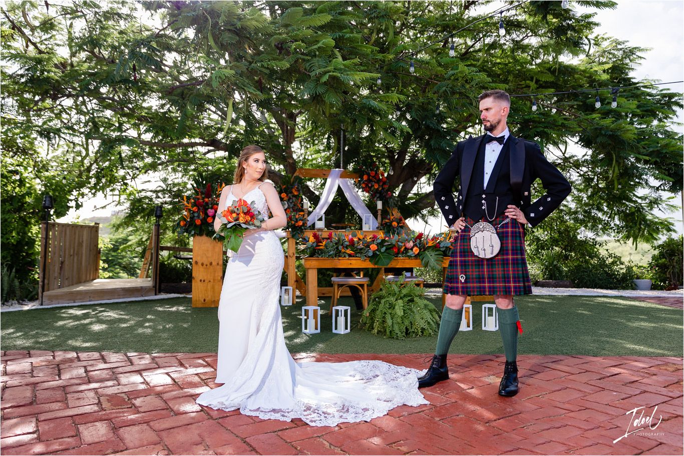 Outdoor wedding ceremony setup with wooden arch, lanterns, and couple in traditional Scottish and bridal attire.