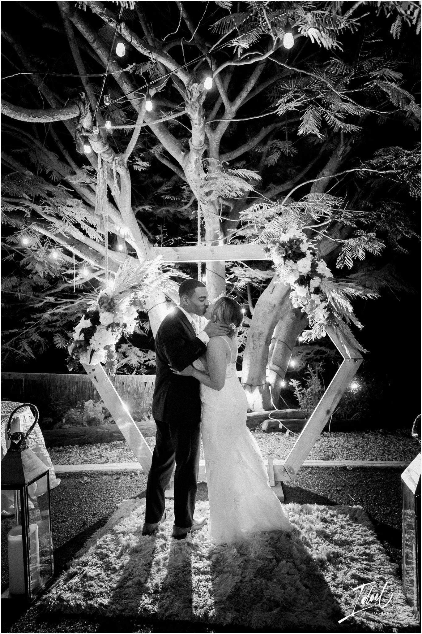 A wedding ceremony under a hexagonal arch with string lights in a garden setting at night.