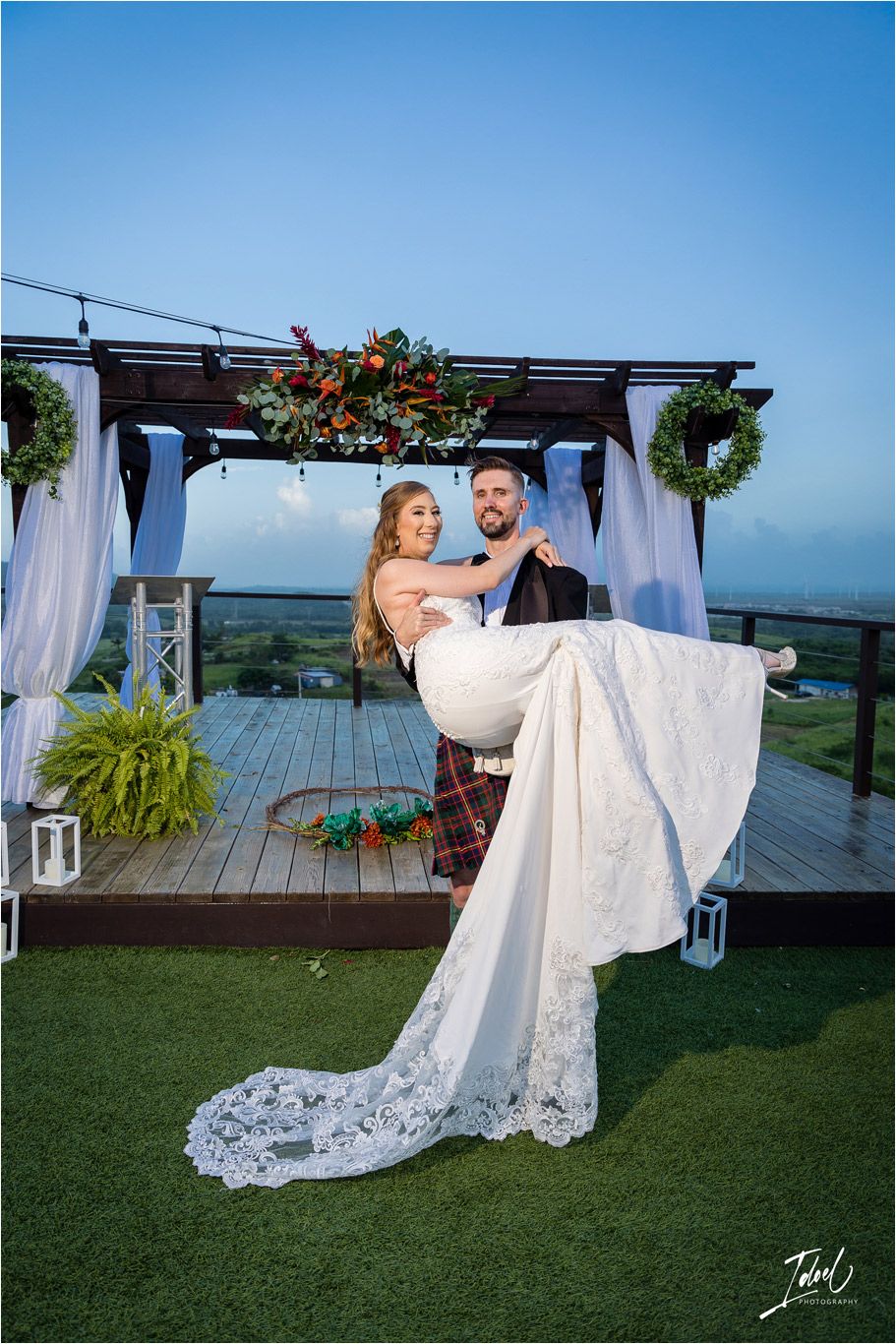 Traditional wedding lift pose under decorated pergola with scenic mountain backdrop.