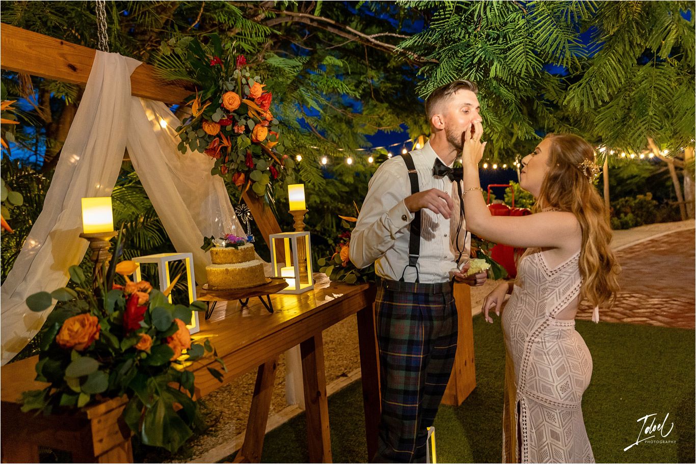 Couple playfully feeds each other cake at outdoor reception with string lights.